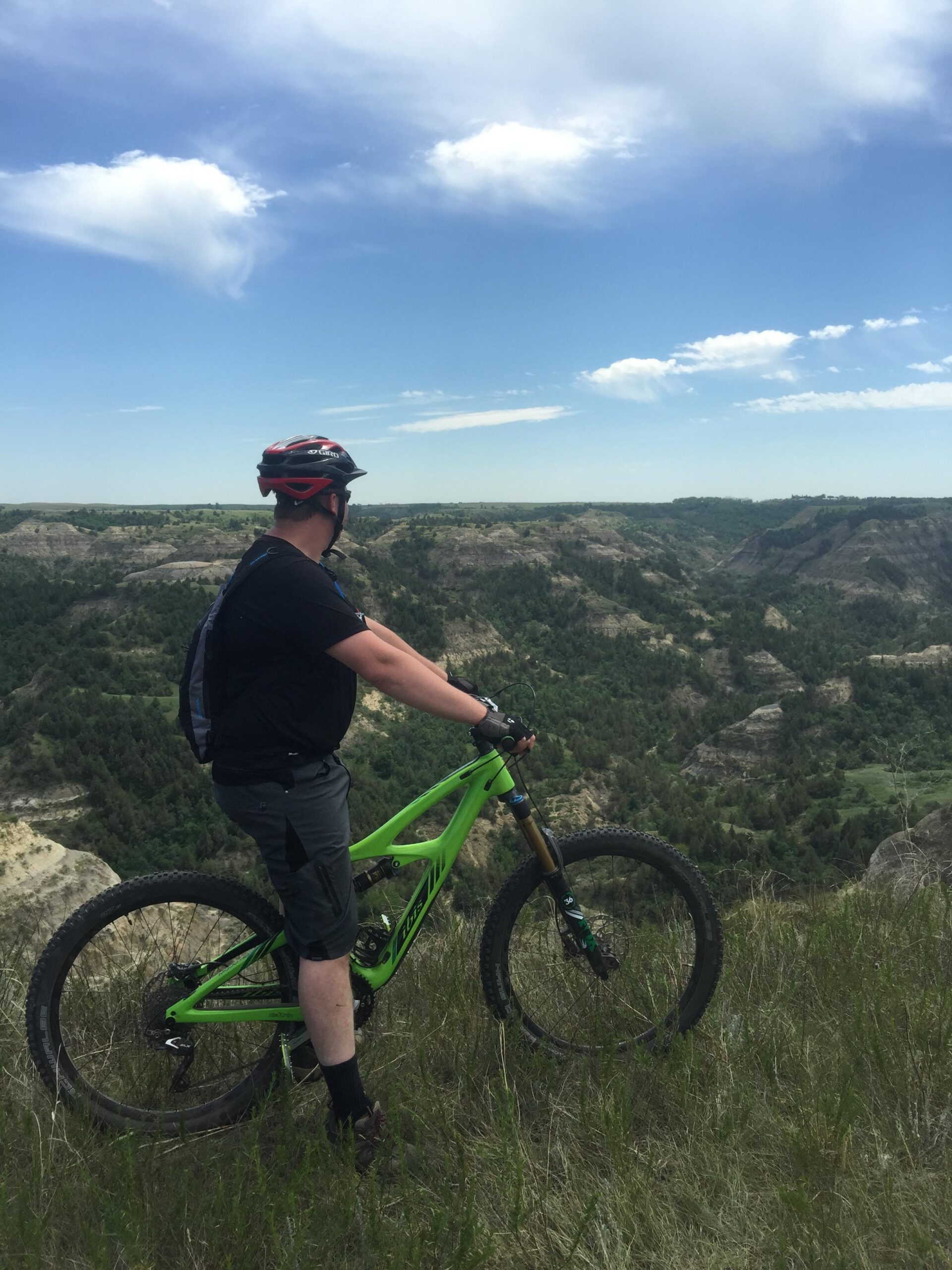 Ibis Mojo HD3: A person wearing a helmet and athletic attire stands beside a green mountain bike, looking out over a scenic landscape with rolling hills and a blue sky dotted with clouds. The surroundings are lush with vegetation, creating a panoramic view of nature.
