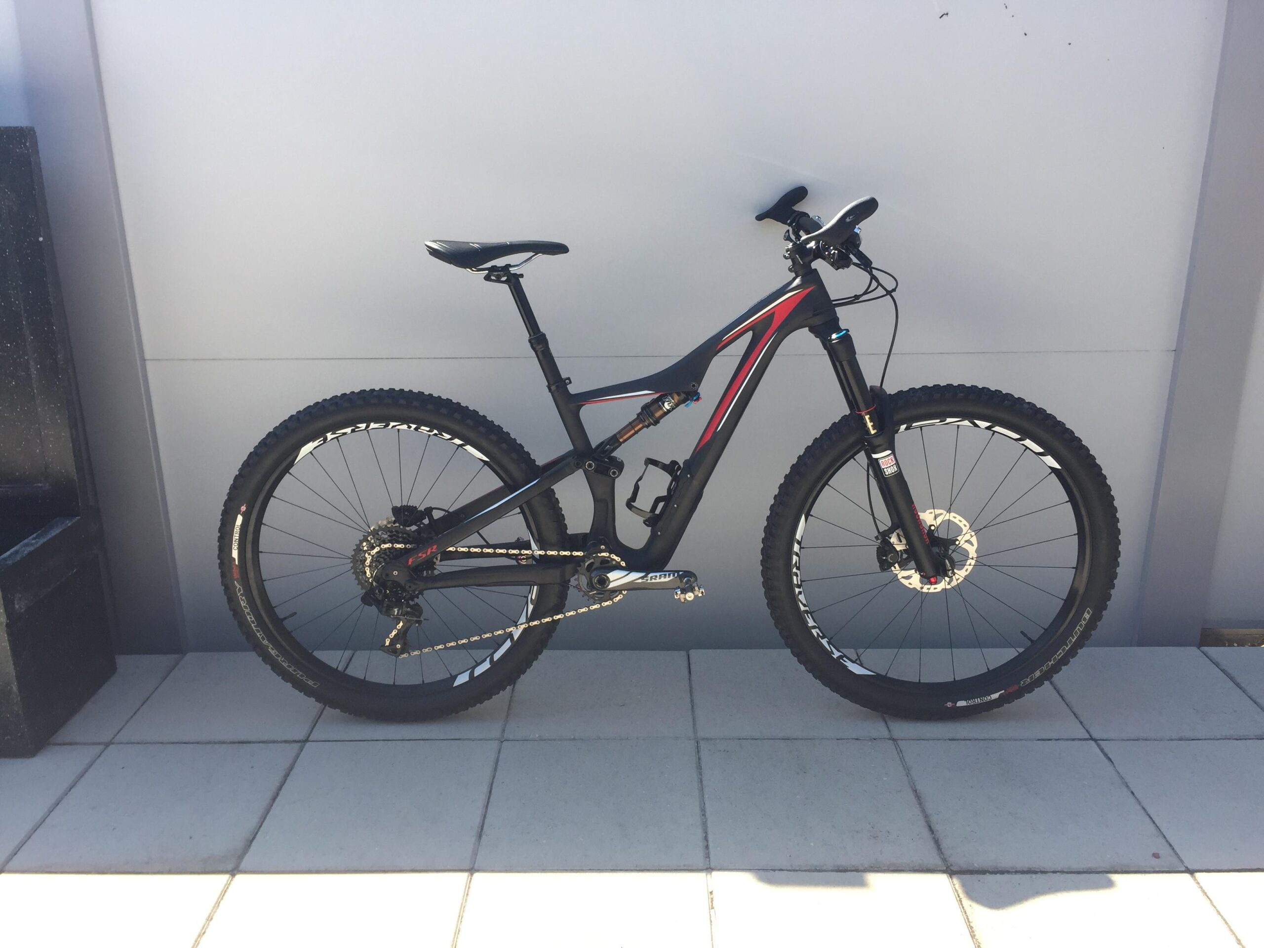 Specialized Stumpjumper FSR Expert Carbon Evo 650B: A black and red mountain bike leaning against a gray wall, with visible thick tires and a suspension system. The bike is positioned on a tiled surface with sunlight illuminating it.