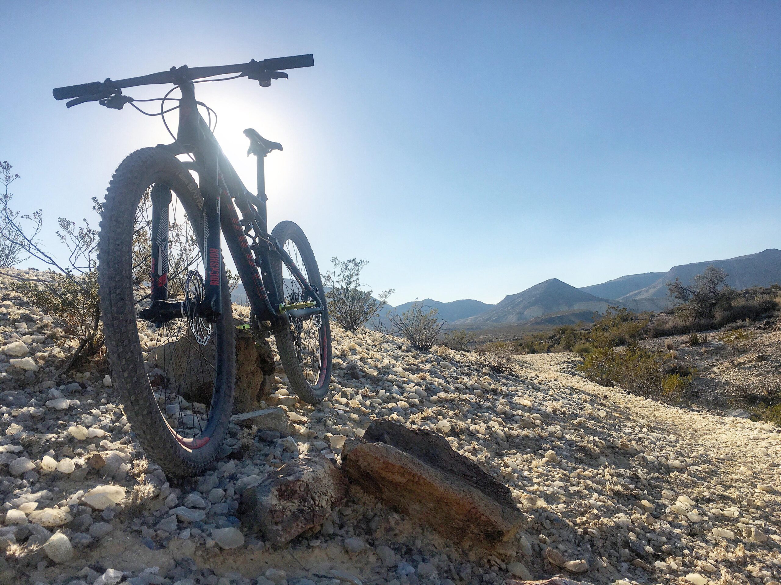 A mountain bike parked on rocky terrain, with the sun positioned behind it, creating a silhouette effect. In the background, rolling hills and sparse vegetation are visible under a clear blue sky. Big Bend Ranch State Park mountain bike trail.