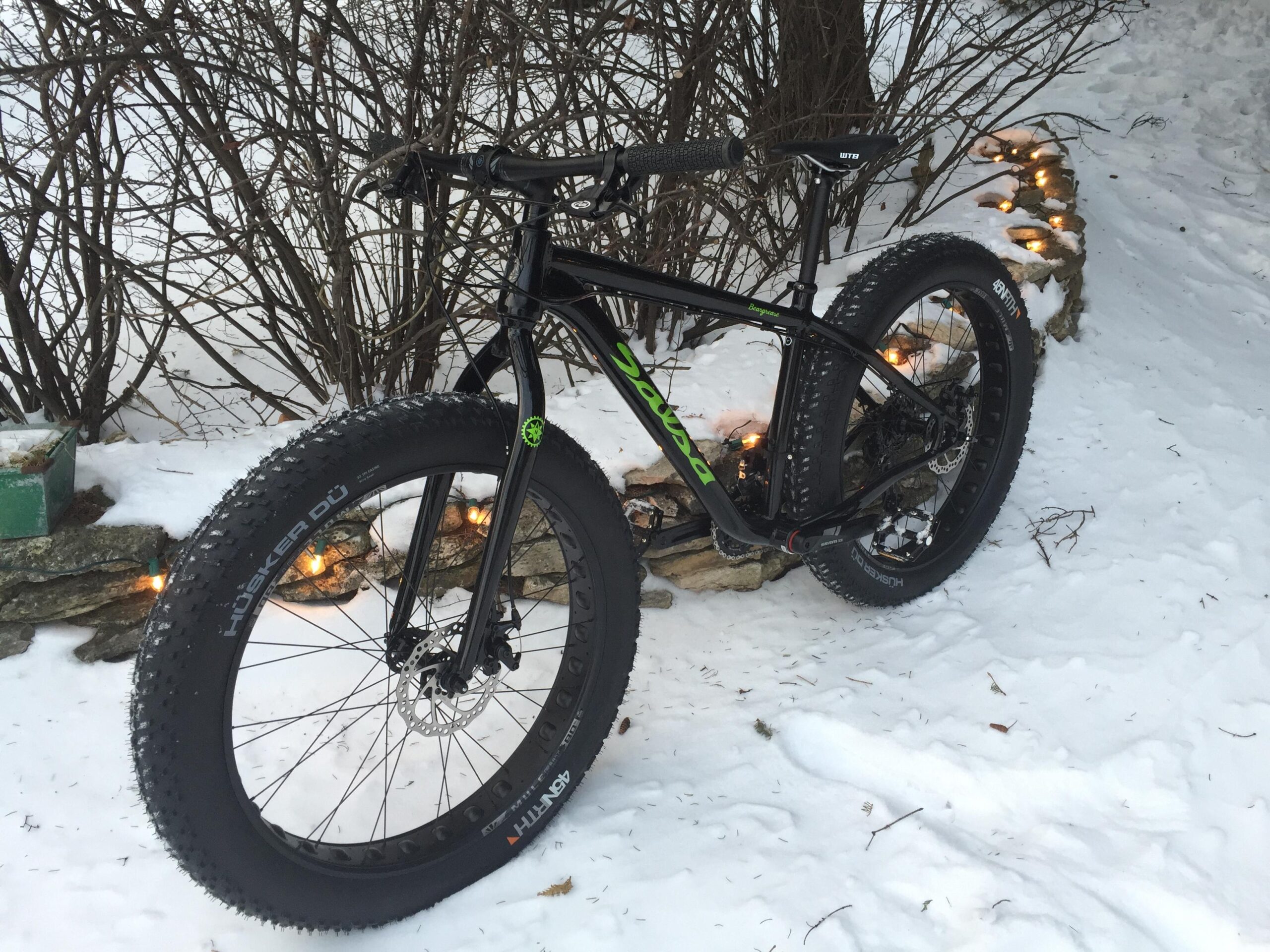 Salsa Beargrease: A black fat bike parked on snow-covered ground, featuring wide tires with a knobby tread pattern, leaning against a snow-dusted stone wall. In the background, there are bare bushes and a soft glow from lights intertwined with the stones.