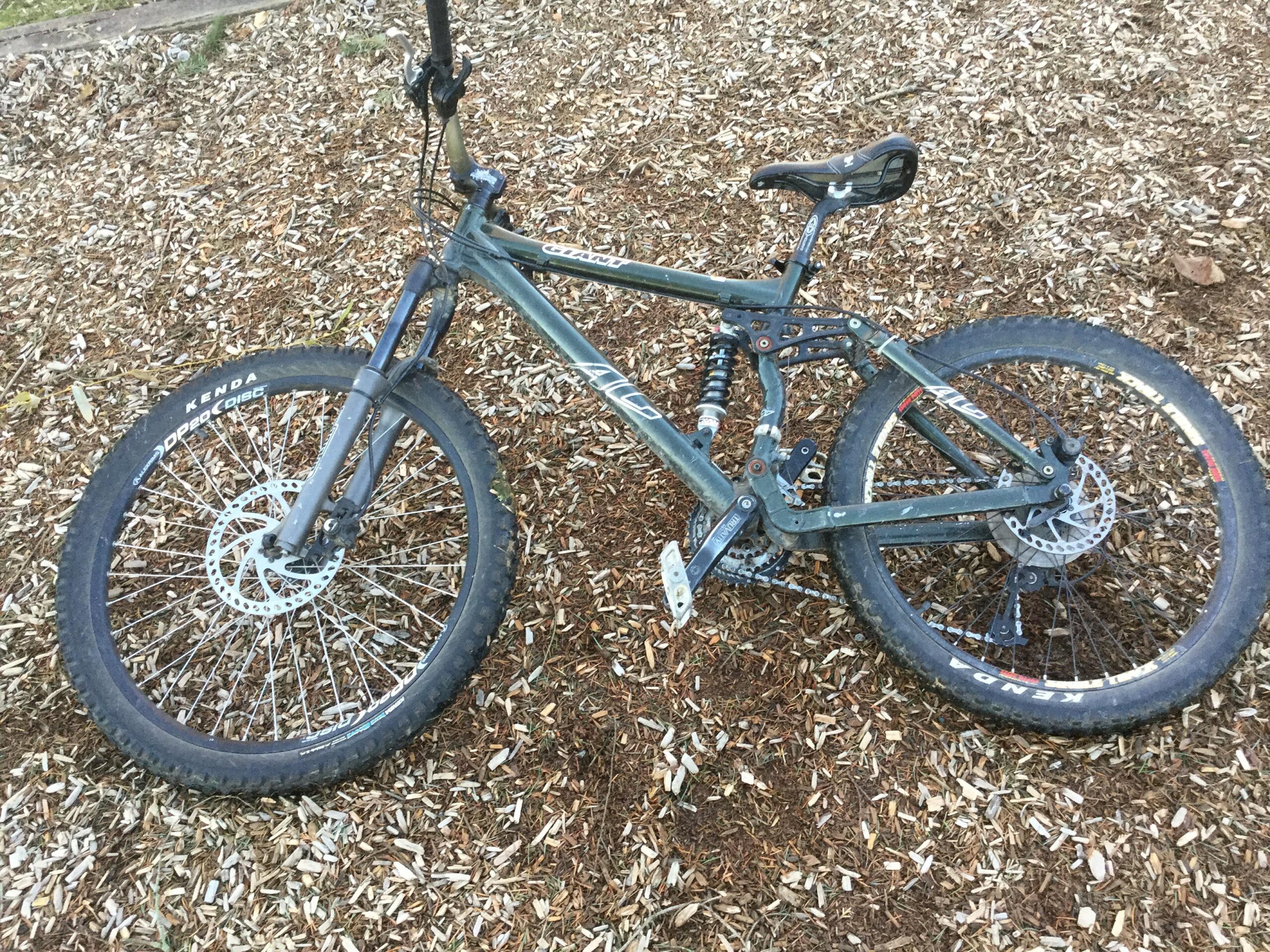 Giant AC: A mountain bike lying on a bed of wood chips, featuring a gray frame, front suspension forks, and wide tires with Kenda branding. The bike is positioned at an angle, showcasing its disc brakes and gear components.