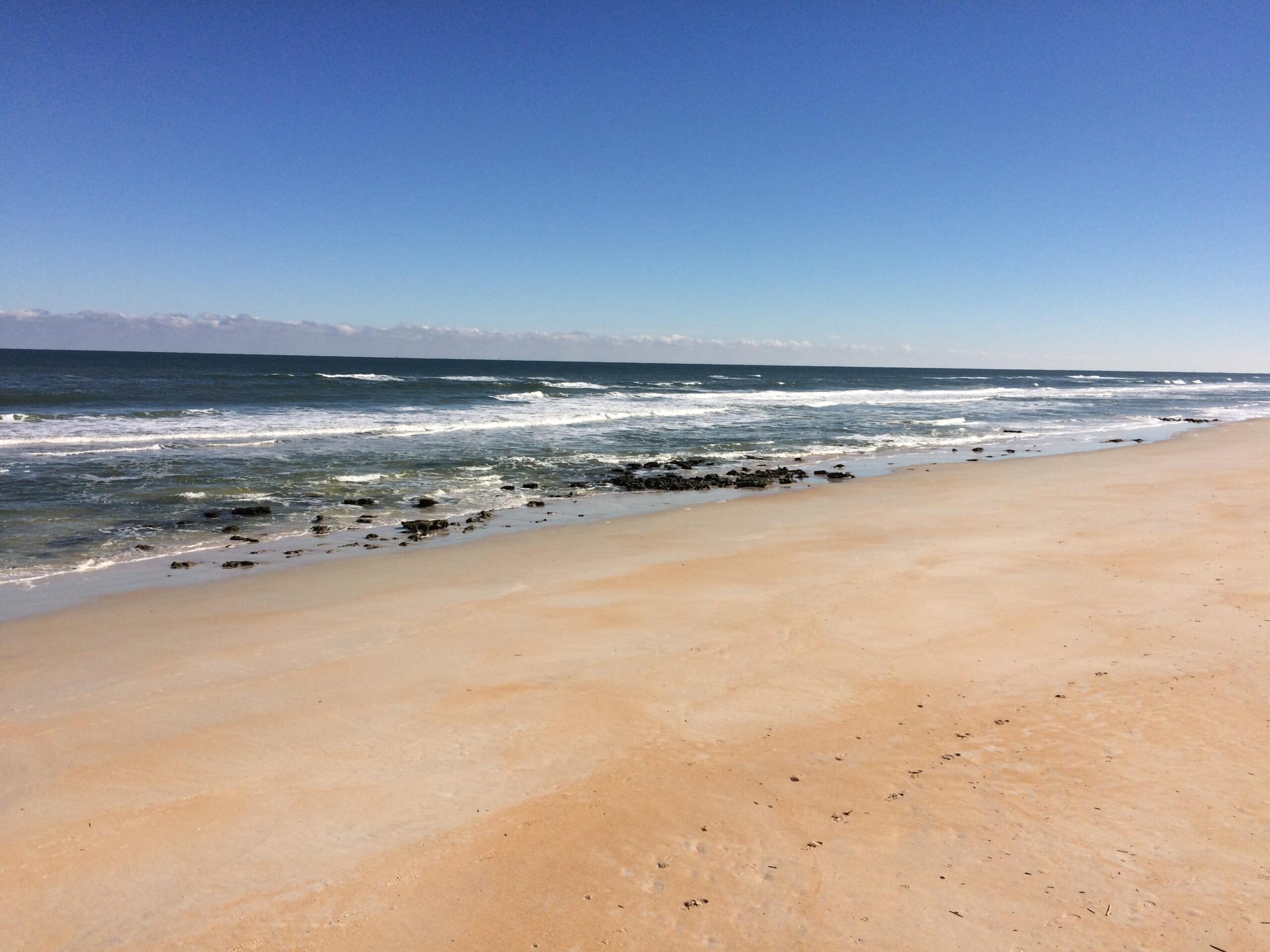 A tranquil beach scene with soft, golden sand leading to gentle waves lapping at the shore. The ocean stretches into the distance under a clear blue sky, with a few wispy clouds on the horizon. Rocky formations are visible near the water's edge. Mala Compra mountain bike trail.