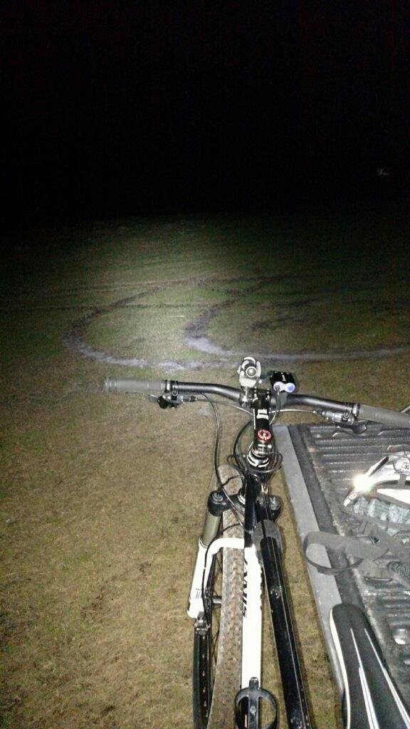 A mountain bike is parked on a grassy area at night, with its front wheel angled towards the ground. The bike's headlight illuminates the ground, revealing tire tracks in a swirling pattern nearby. The background is dark, with minimal visibility. Governor's Creek mountain bike trail.