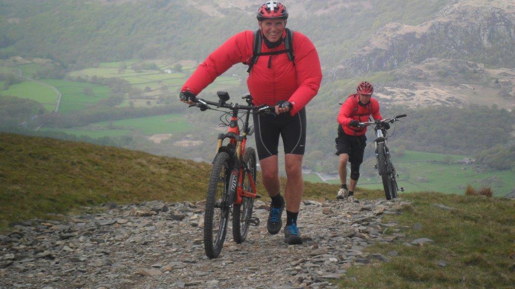 Trek Remedy 9.7 27.5/650b: Two mountain bikers ascending a rocky trail, wearing bright red jackets and helmets. One cyclist is in the foreground smiling as he pushes his bike uphill, while the other cyclist trails behind. Below, a scenic view of rolling green hills and fields is visible, under a misty sky.