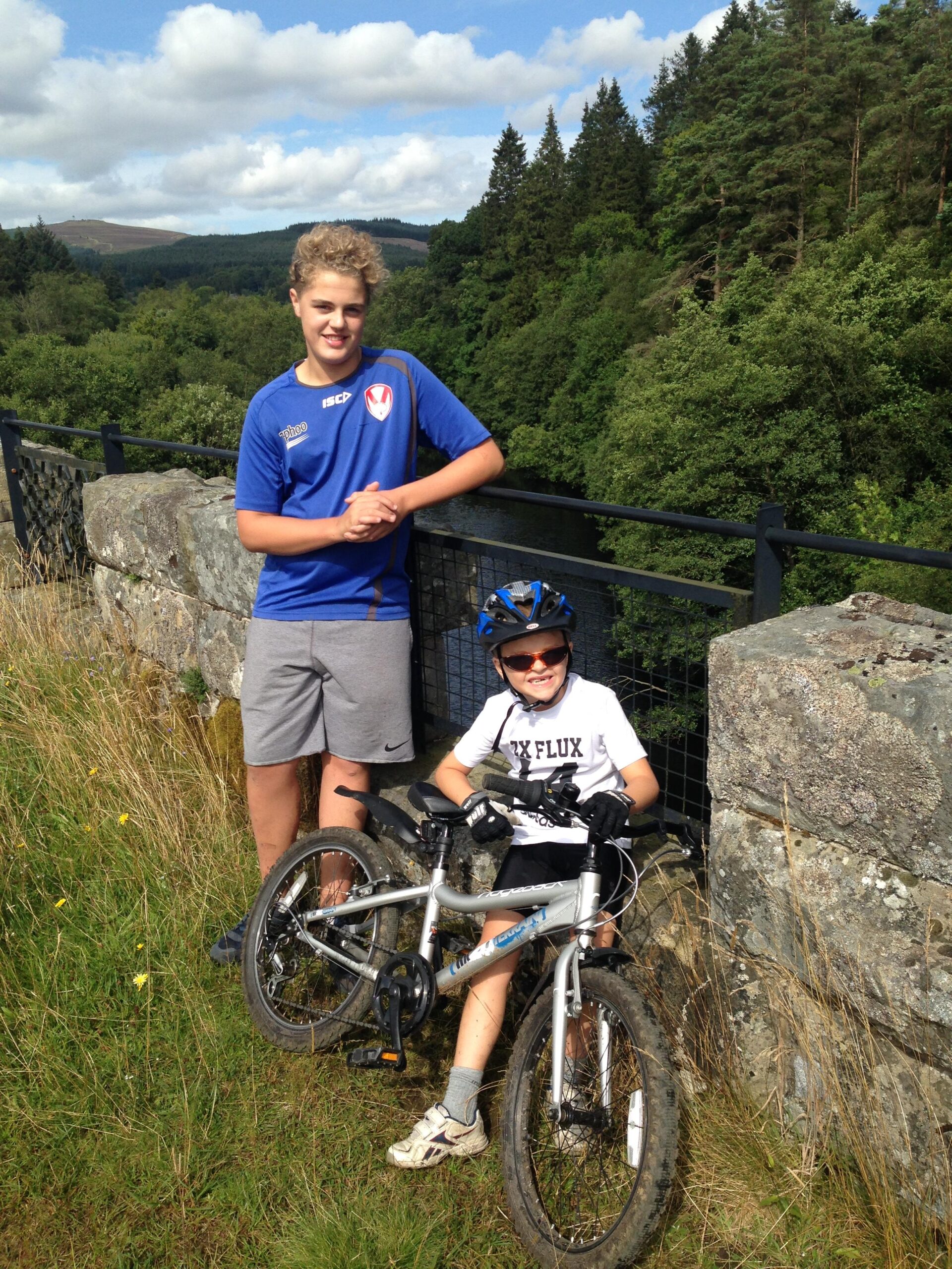 Trek Remedy 9.7 27.5/650b: Two boys are posing together next to a bicycle on a grassy area beside a stone wall. The older boy stands with his arms crossed, wearing a blue sports shirt and gray shorts, while the younger boy sits on a silver bike wearing sunglasses and a white t-shirt with black lettering. The background features a lush forest and hills under a partly cloudy sky.