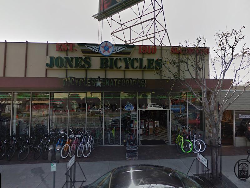 Front view of Jones Bicycles storefront, featuring bikes and skateboards on display. The sign prominently displays "JONES BICYCLES" and "Established 1910." The shop has a checkered floor visible through the windows and a mix of bicycles lined up outside.
