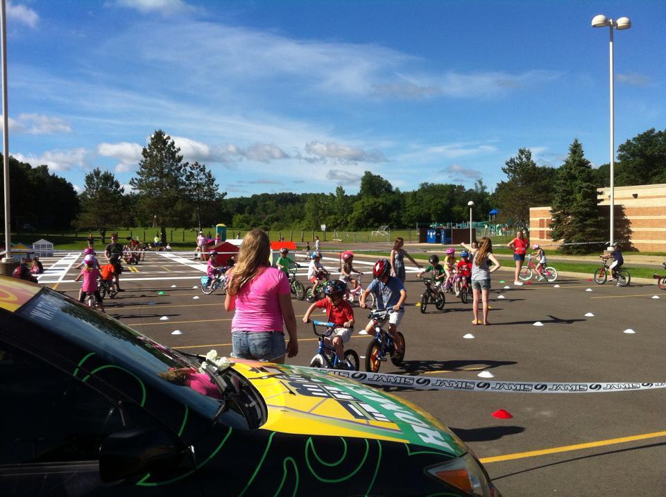 A sunny day at a parking lot where children are riding bicycles on a marked course, attended by adults. The scene includes various kids wearing helmets, some in colorful bikes, while others are being guided by adults. There are traffic cones and markers set up for a bike safety event, with a school building and playground visible in the background.