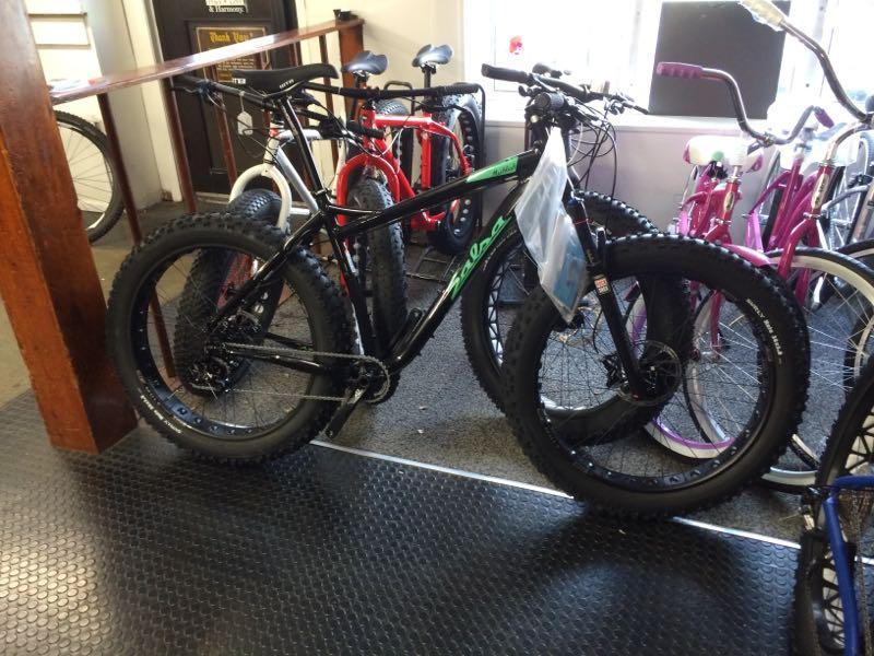 A black fat tire bike with green accents is displayed in a bike shop, surrounded by various other bicycles of different colors, including red and pink. The bike features wide, knobby tires and a price tag attached. The shop has a wooden railing and tile flooring, with natural light coming through the windows.
