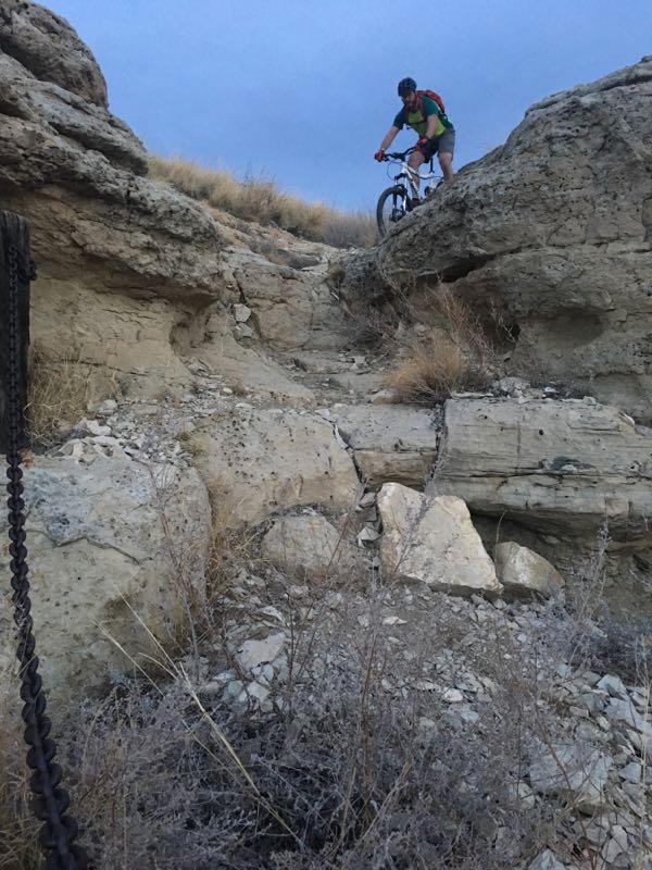 A mountain biker navigating a rocky trail, skillfully balancing on uneven terrain surrounded by dry grasses and rocky outcrops under an overcast sky. Steep Tech mountain bike trail.