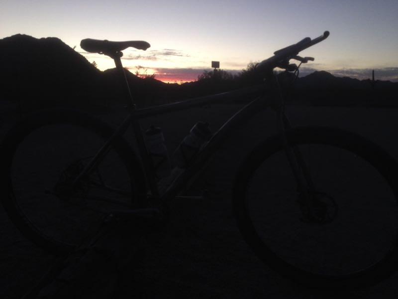 Silhouette of a bicycle against a sunset backdrop, featuring mountains and a colorful sky transitioning from orange to dark blue. Two water bottles are mounted on the bike frame. The image captures a tranquil outdoor scene during dusk. San Tan mountain bike trail.