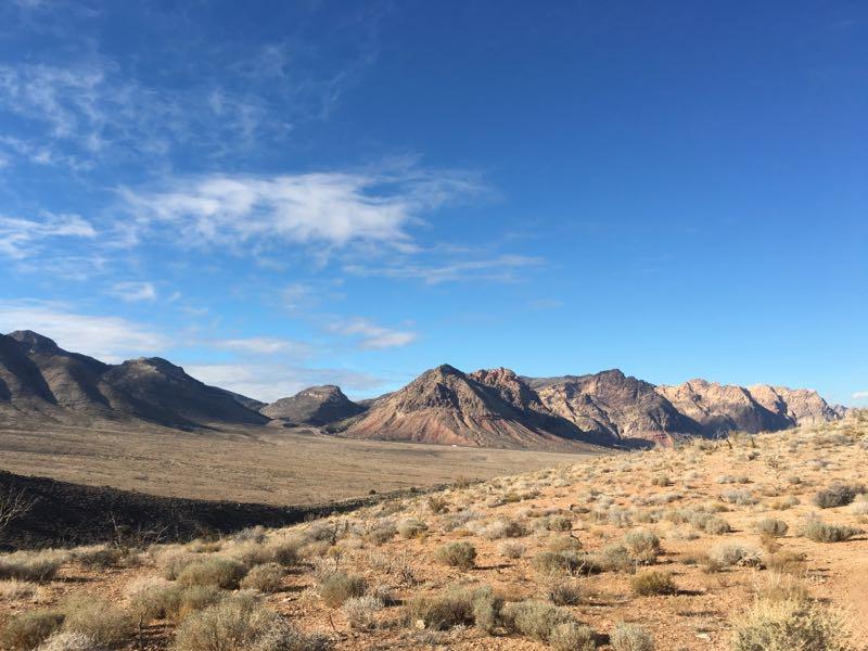 A wide landscape view of mountainous terrain under a blue sky, featuring rocky outcrops and sparse vegetation. The foreground shows a barren area with scattered shrubs, leading up to distinct mountain peaks in the background. Badger Pass Loop mountain bike trail.