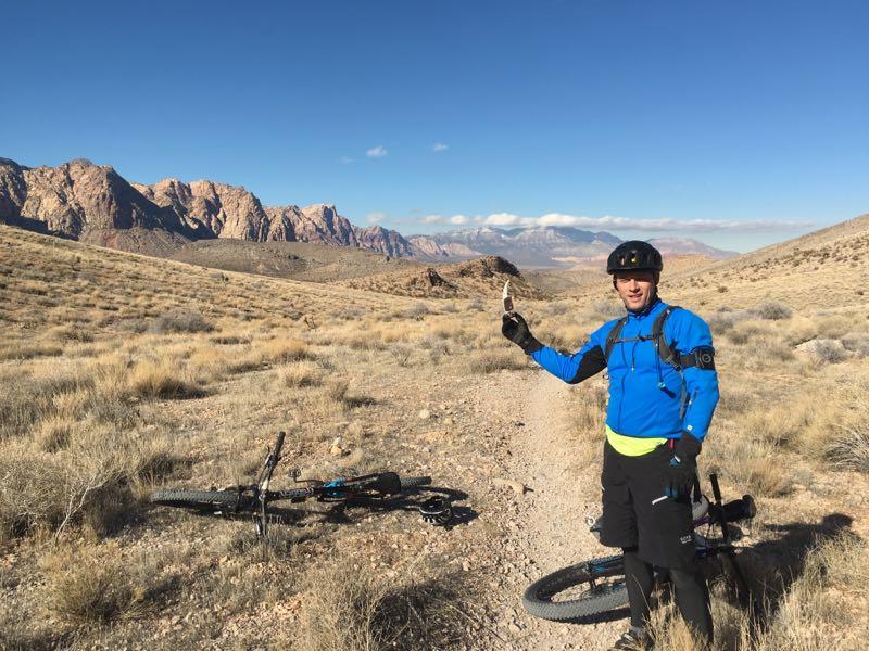 A mountain biker in a blue jacket and black helmet stands on a dirt trail, holding up a tool or snack, against a backdrop of rocky mountains and clear blue skies. Two bicycles lie on the ground nearby, surrounded by dry grass and shrubs. Badger Pass Loop mountain bike trail.