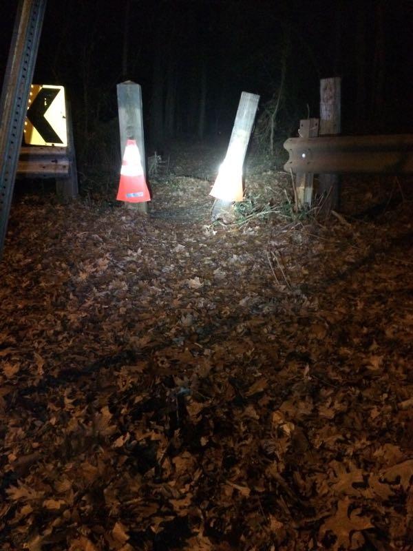 A dimly lit path through a forest at night, featuring two orange traffic cones marking the way. The ground is covered in fallen leaves, and a caution sign indicating a curve is visible on the left. The scene appears quiet and secluded. Trails seperated by streets mountain bike trail.