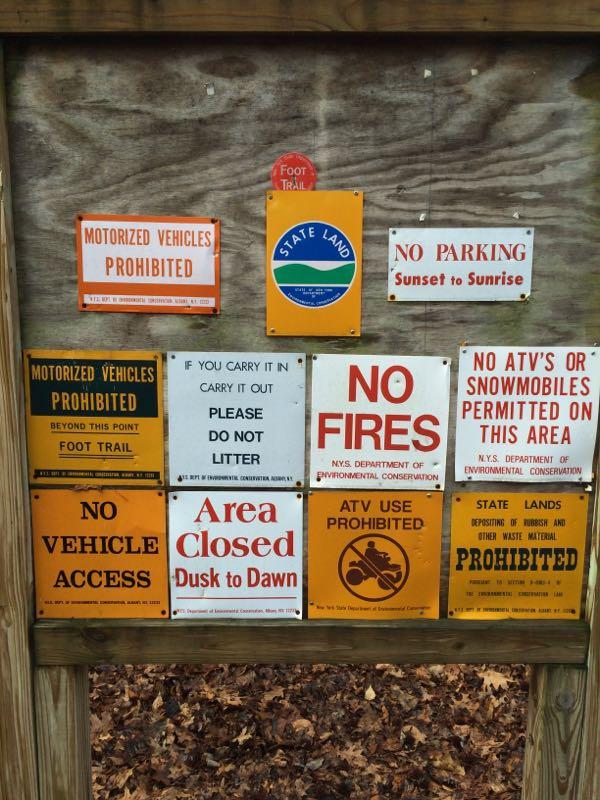 A collection of signs mounted on a wooden board, displaying various regulations for a public outdoor area. The signs include warnings against motorized vehicles, littering, and fires, along with instructions for foot trail access, parking restrictions, and prohibitions on ATV and snowmobile use. The "state land" sign is prominently featured amid safety and access rules. The background includes leaves on the ground, indicating it’s an outdoor setting. Long Pond mountain bike trail.