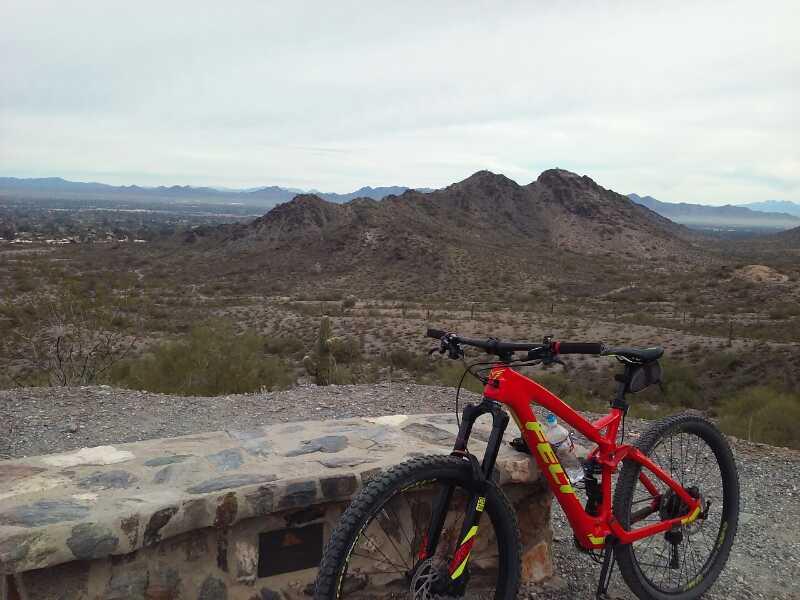 A red mountain bike rests on a stone wall overlooking a desert landscape with rolling hills and distant mountains under a cloudy sky. Trail #100 mountain bike trail.