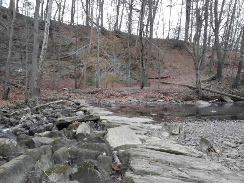 A rocky streambank with a gentle flow of water, surrounded by trees with bare branches and fallen leaves. A steep, rocky hillside is visible in the background, creating a natural setting in a wooded area during late autumn. Smedley park mountain bike trail.