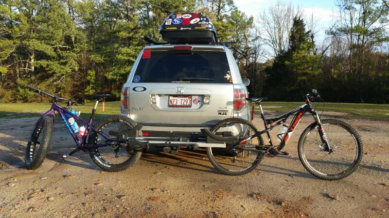 A silver SUV parked on a gravel surface, equipped with a bike rack holding two mountain bikes—one purple and one black/red. The background features trees, suggesting a natural outdoor setting ideal for biking. Clinton Nature Preserve mountain bike trail.