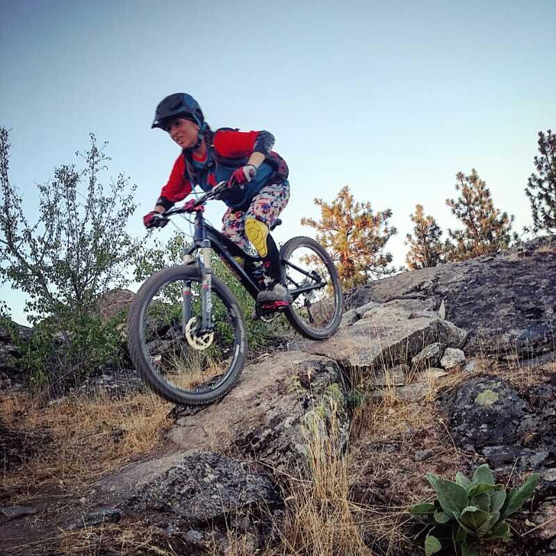 A person riding a mountain bike along a rocky trail with vegetation in the background. The individual is wearing a helmet and colorful cycling gear, demonstrating an energetic riding posture as they navigate the terrain. The setting appears to be outdoors in a natural landscape. Beacon Hill mountain bike trail.