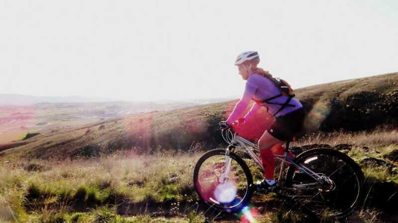 A mountain biker in a purple long-sleeve shirt and helmet rides along a grassy trail, with a panoramic view of rolling hills in the background. The sun shines brightly, creating a lens flare effect in the foreground. Saltese Conservation Area mountain bike trail.