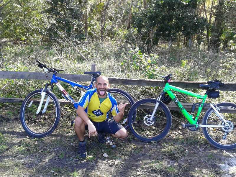 A man wearing a bright yellow and blue cycling jersey kneels in front of two mountain bikes, smiling and sticking out his tongue. The bikes are positioned next to a wooden fence in a natural outdoor setting with greenery in the background. The scene conveys a sense of joy and enthusiasm for cycling. Soldier Creek Park mountain bike trail.