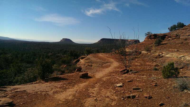A rugged dirt path winds through a landscape of red rock formations and sparse vegetation, with distant mesas under a clear blue sky. The scene captures the natural beauty of a desert environment. Mescal Trail mountain bike trail.