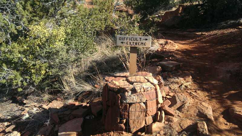Wooden sign pointing in two directions, labeled "Difficult" and "Extreme," sits atop a stone marker on a rugged hiking trail surrounded by vegetation. Mescal Trail mountain bike trail.