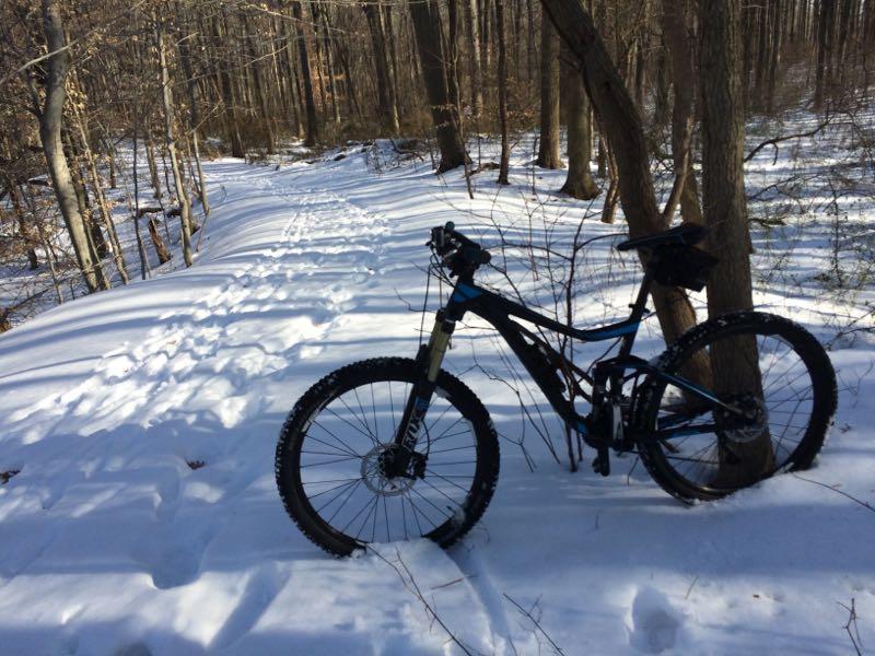 A mountain bike resting against a tree on a snow-covered trail in a forest. Footprints are visible in the snow, leading up the winding path through the wooded area. Wolfes Pond park mountain bike trail.