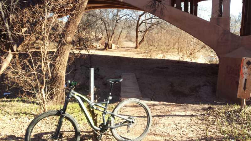 A mountain bike resting on the ground beside a dirt path, with a bridge overhead and sparse trees in the background. The environment appears dry, hinting at a natural recreational area. Wee-chi-ta mountain bike trail.