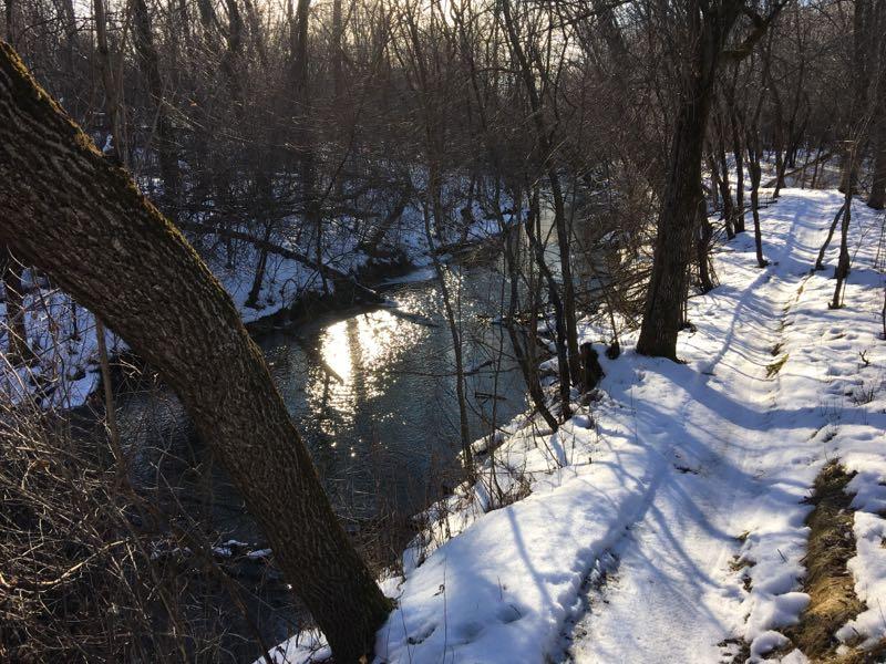 A tranquil winter scene featuring a narrow creek surrounded by bare trees. The water reflects sunlight, creating a shimmering effect on its surface. A snow-covered path follows the creek's edge, and the ground is partially blanketed in snow, suggesting a peaceful, quiet natural setting. Bertram Chain of Lakes Trail mountain bike trail.