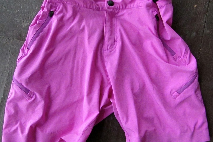 A pair of vibrant pink shorts laid flat on a wooden surface, featuring a button and zipper closure, an adjustable waistband with straps, and two side pockets.
