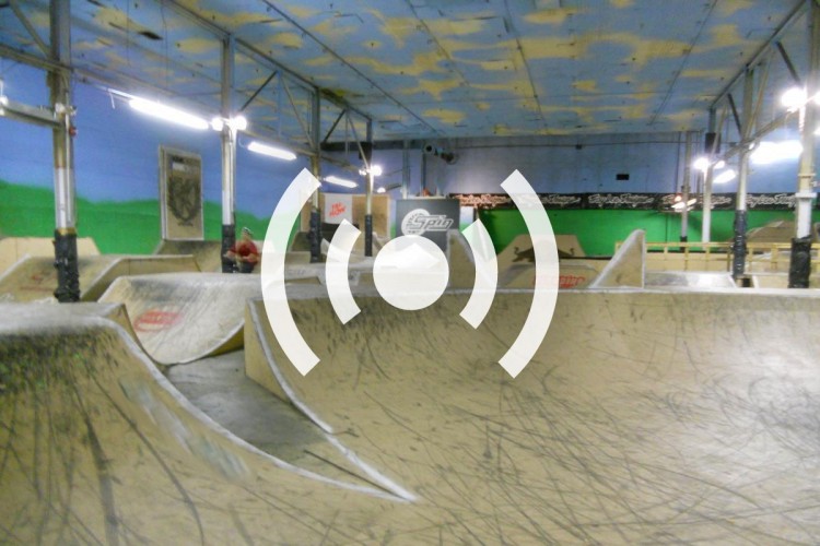 An indoor skate park featuring a series of ramps and bowls, with graffiti-style art on the walls. The ceiling is painted blue with yellow clouds. Overhead lights illuminate the space, and a few skaters can be seen in the background.