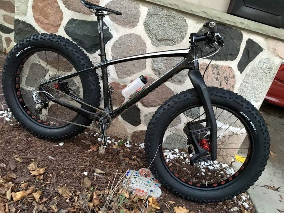 Borealis Yampa: A black fat bike leaning against a stone wall, featuring wide tires designed for stability on soft or uneven terrain. The bike has a sleek frame and is equipped with a water bottle mounted on the side. Surrounding the bike are fallen leaves and white pebbles.