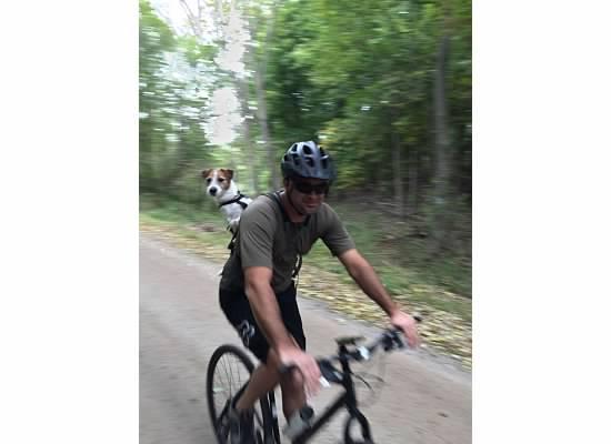 A person wearing a helmet and sunglasses rides a bicycle along a dirt path, carrying a small dog in a backpack-like harness on their back, surrounded by trees. Anderson Park mountain bike trail.