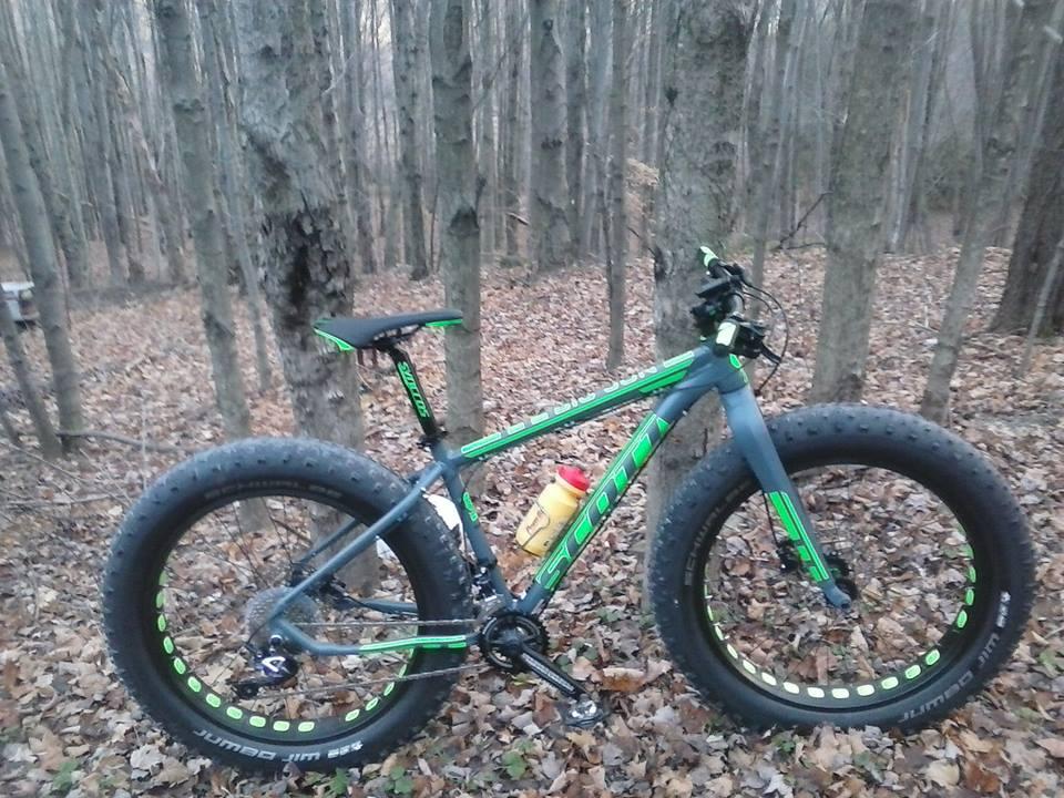 Scott Big Jon: A fat tire mountain bike with a gray and green frame is parked among trees in a wooded area covered with fallen leaves. The bike features oversized tires and a water bottle attached to the frame, showcasing its design for rugged terrain.