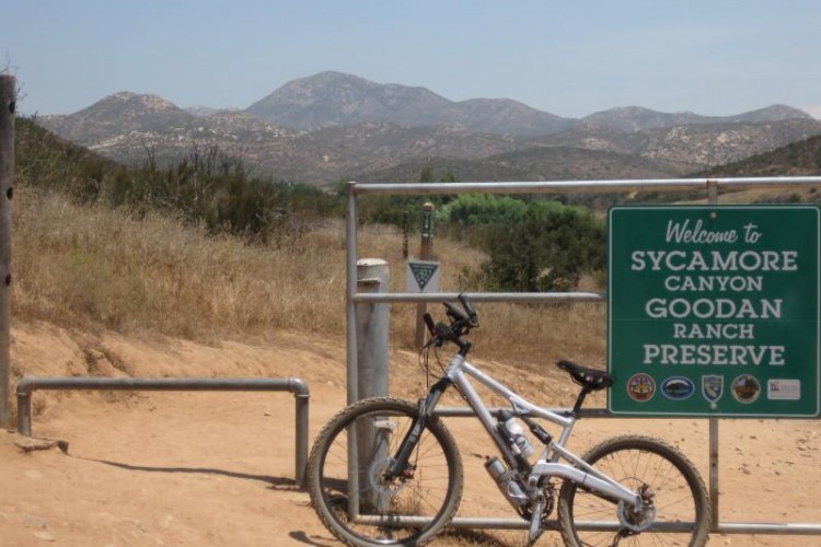 Alt text: A white mountain bike leans against a metal gate with a green sign that reads "Welcome to Sycamore Canyon Goodan Ranch Preserve." In the background, rolling hills and mountains can be seen under a clear blue sky. The landscape features dry grass and sparse vegetation.