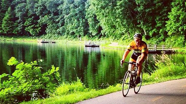 A cyclist in a yellow and black jersey rides along a paved path beside a calm lake, surrounded by lush green trees. The water reflects the vibrant greenery, creating a picturesque and serene outdoor scene.