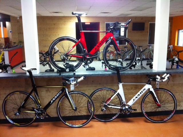 A display of three Jamis bicycles arranged on a showroom floor. The top bike is a sleek red model, while the two bikes below are black and white models. The background features a wooden wall and additional bicycles lined up, suggesting a bike shop environment.