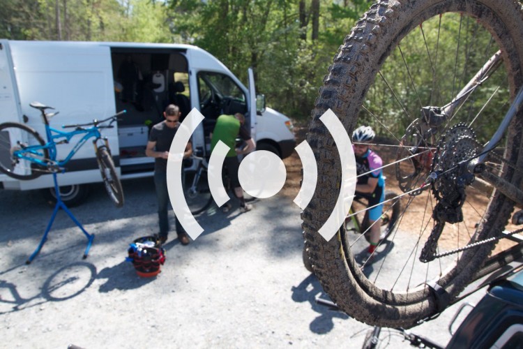 A group of cyclists is gathered around a white van in a wooded area, with bicycles being repaired and maintained. One bike is mounted on a repair stand, while two cyclists are engaged in maintenance work. The scene captures the camaraderie of outdoor cycling activities, surrounded by trees and gravel.