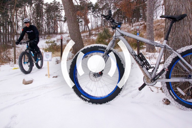 A rider on a fat bike navigating a snowy trail surrounded by trees, with a sign indicating trail conditions in the background. The foreground features a close-up of the fat bike's large tires, emphasizing the winter cycling environment.