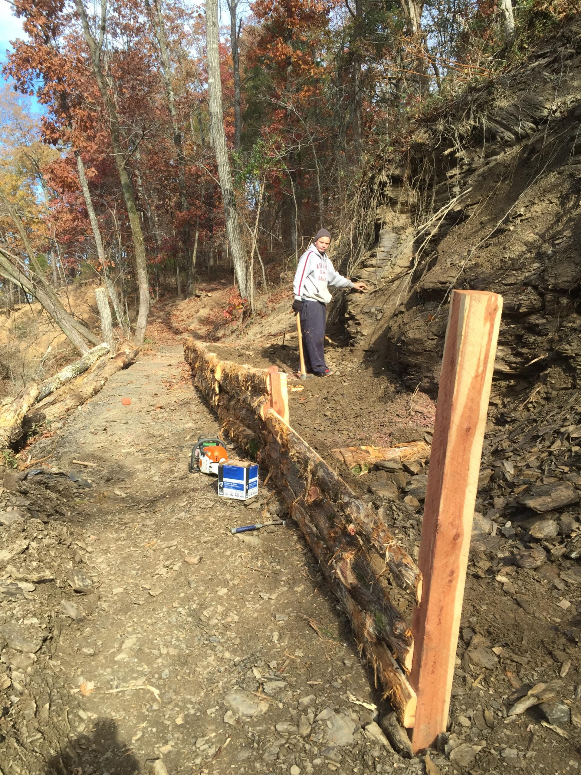 A person stands beside a partially constructed wooden barrier on a dirt path in a forested area. The landscape features autumn-colored trees in the background. The individual is wearing a hoodie and sweatpants and is holding a tool while working on the project. Nearby, there is a chainsaw and a box on the ground, indicating ongoing construction work. Spadra Creek Nature Trail mountain bike trail.