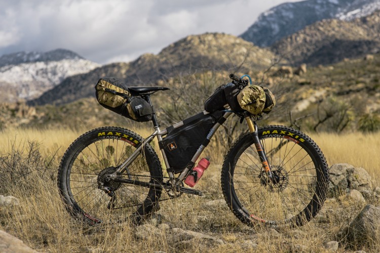 A rugged mountain bike with various packs and gear attached stands on rocky terrain, surrounded by dry grass and hills in the background. The bike features wide tires suitable for off-road conditions, and its frame is made of a lightweight material. The scenery includes distant mountains partially covered in snow under a cloudy sky.