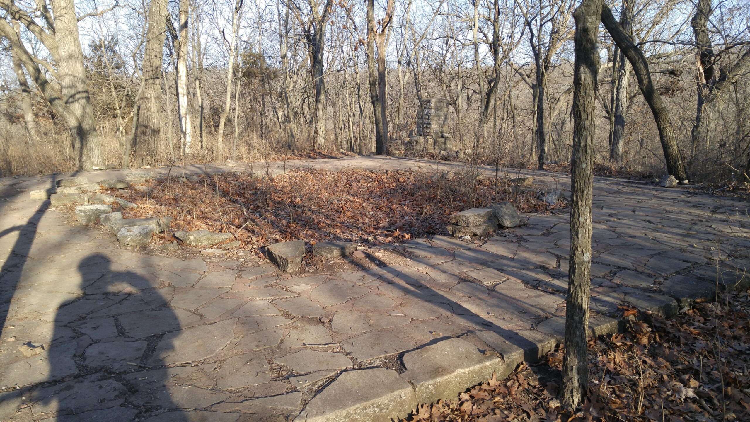 An overgrown stone patio area surrounded by trees, with scattered leaves on the ground and a shadow in the foreground. The site appears abandoned, showing signs of wear and nature reclaiming the space. Swope Park Trail mountain bike trail.