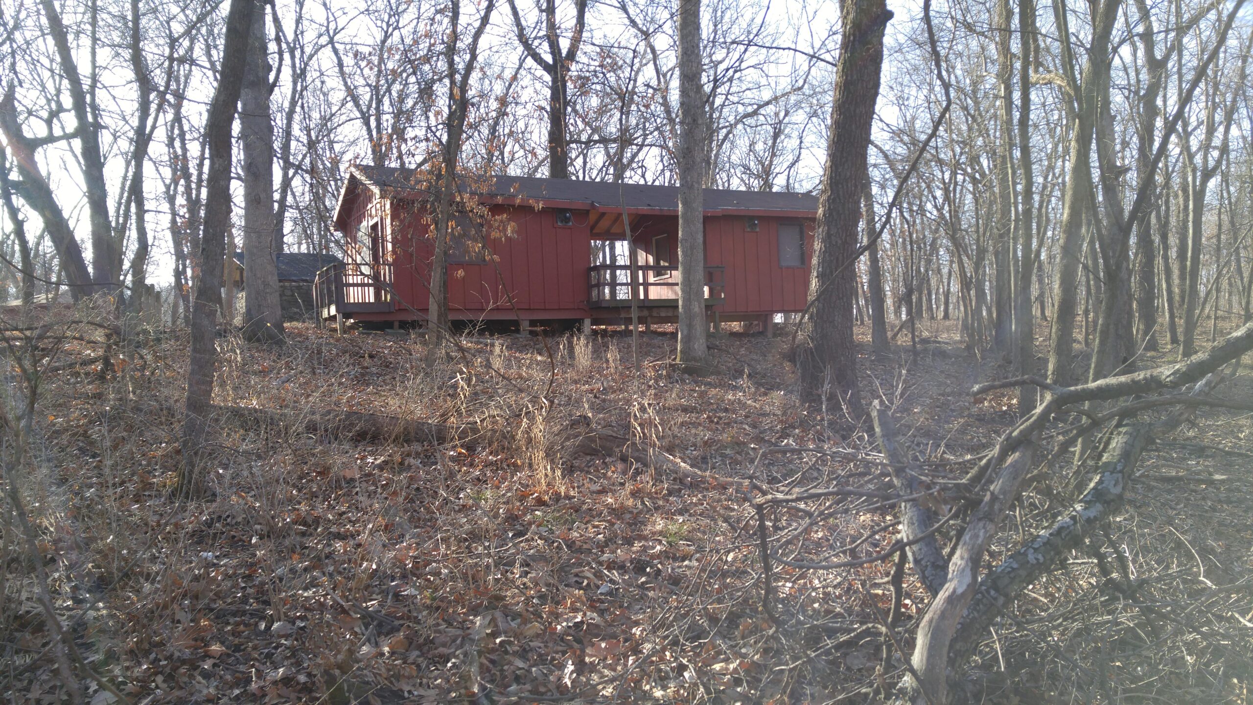 A small red cabin nestled among bare trees on a hillside, surrounded by dry leaves and underbrush. The cabin features a wooden porch and large windows, set against a backdrop of a sparse forest in a late autumn or early winter setting. Swope Park Trail mountain bike trail.