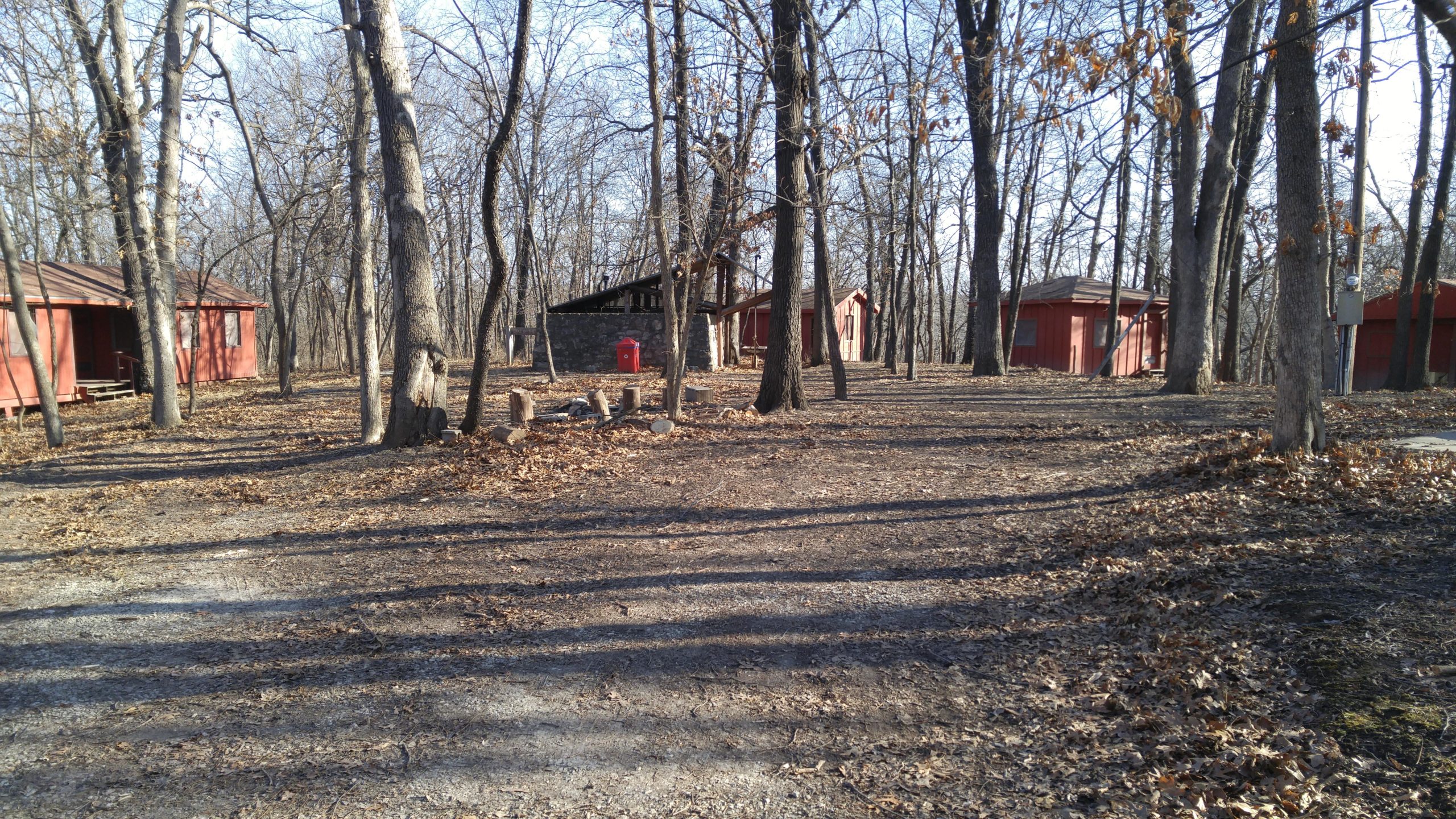 A wooded area featuring several red cabin structures nestled among bare trees, with a gravel path leading through fallen leaves. A stone fireplace and a red mailbox are visible near the cabins, suggesting a rustic retreat or campground setting. Swope Park Trail mountain bike trail.