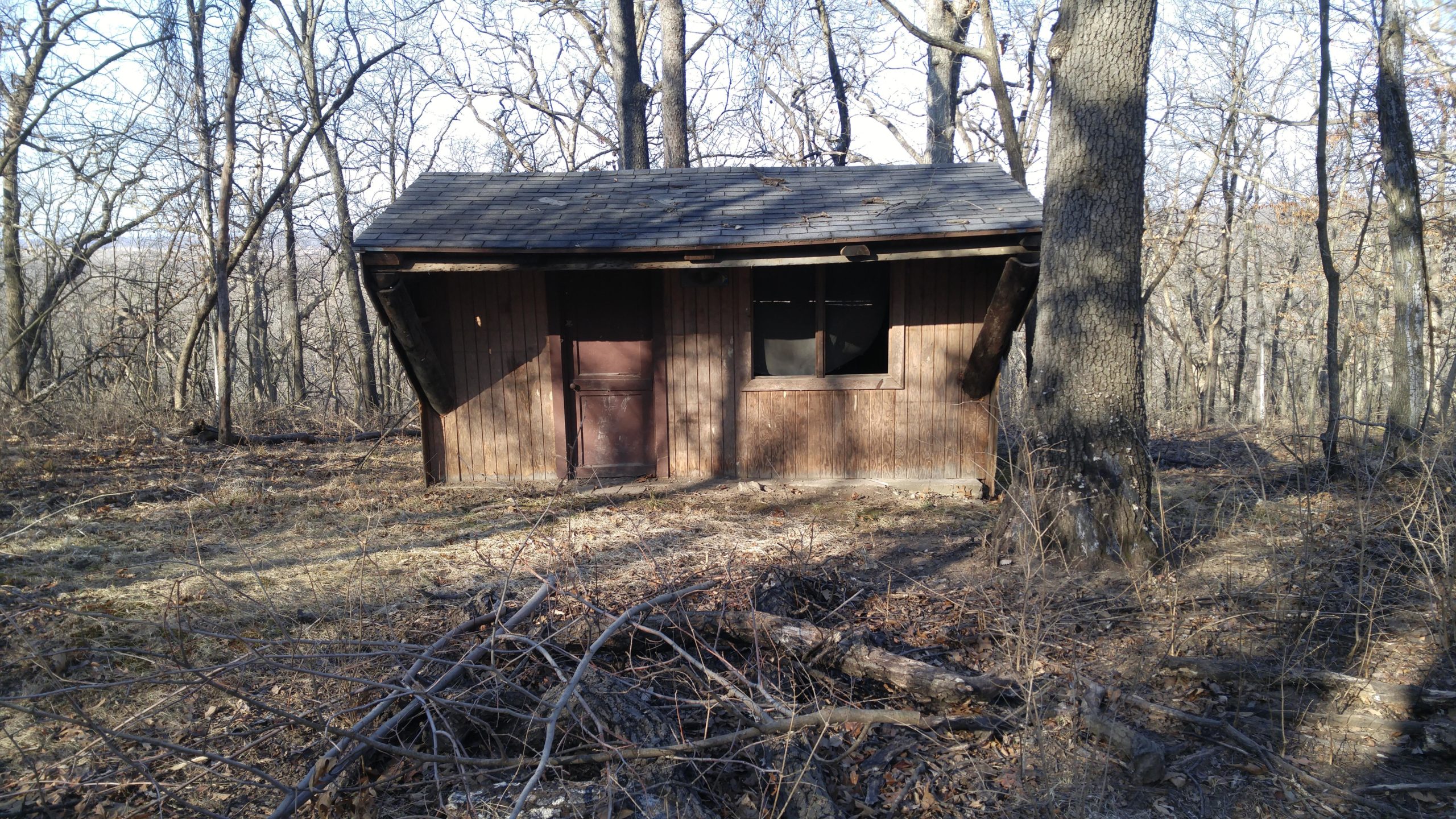 A small, rustic wooden cabin situated in a wooded area with bare trees. The cabin has a slanted roof, a brown door, and a window beside the door. Surrounding the cabin is a patch of dry ground with some fallen branches and sparse vegetation. Swope Park Trail mountain bike trail.