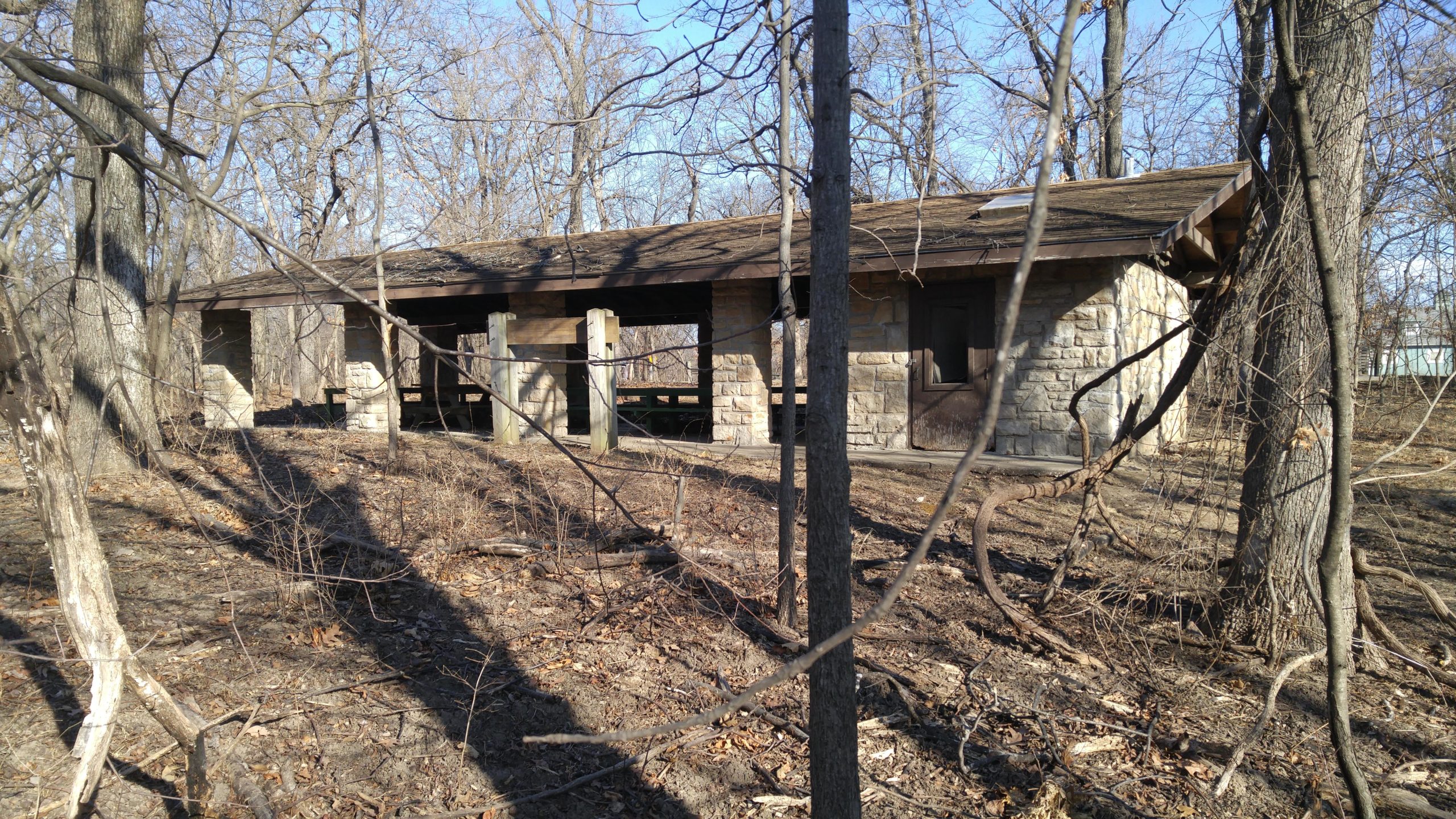 A rustic stone picnic shelter surrounded by bare trees in a wooded area, with sunlight filtering through the branches and shadows cast on the ground, indicating a late fall or early spring setting. Swope Park Trail mountain bike trail.