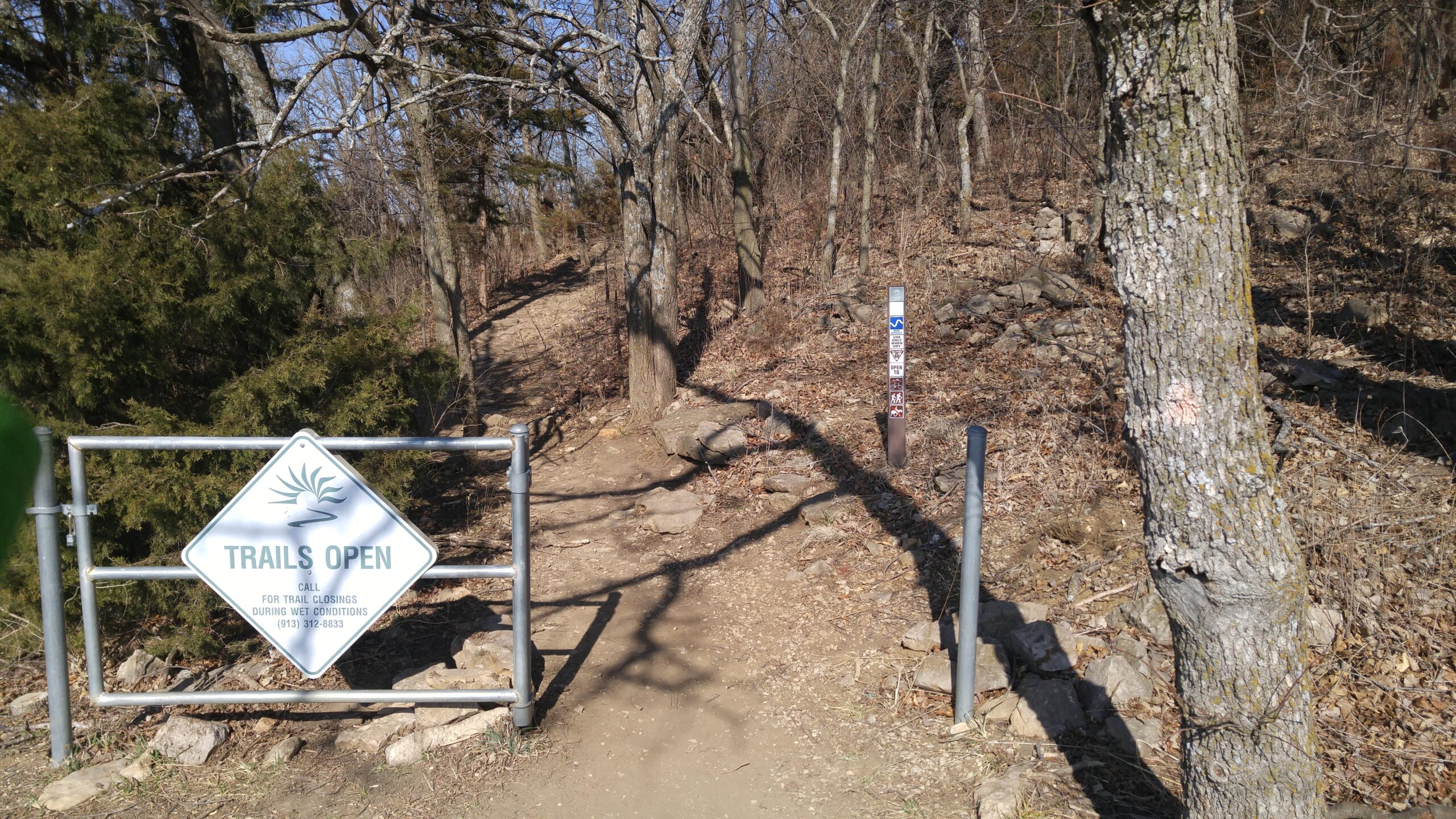 A sign marked "Trails Open" in a diamond shape stands at the entrance to a dirt hiking trail. The path is bordered by trees and rocky terrain, indicating a natural outdoor environment. There is a metal gate leading into the trail, which is partially open, allowing access to the pathway that winds into the woods. Shawnee Mission Park mountain bike trail.