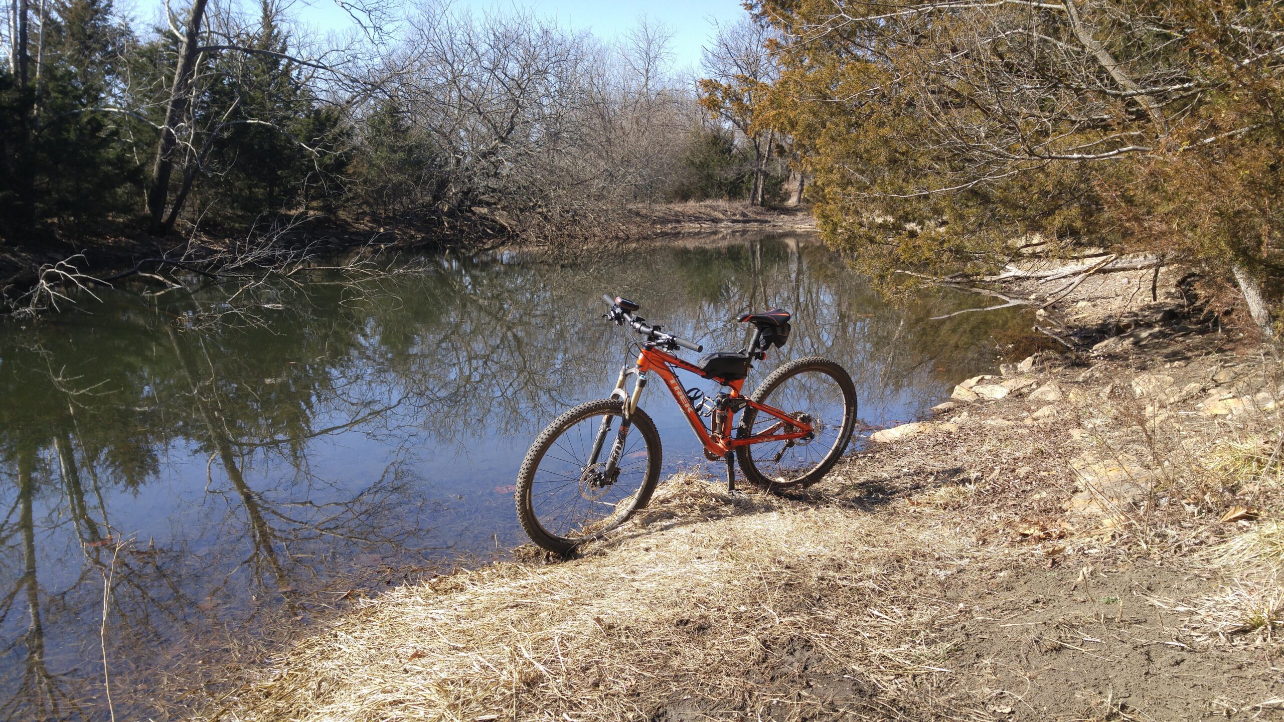 A mountain bike parked on the grassy bank beside a calm river, surrounded by trees and barren branches reflecting on the water's surface. Shawnee Mission Park mountain bike trail.