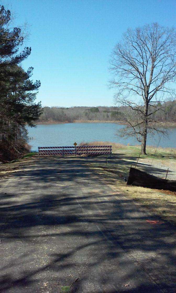 A dirt road leading to a water body is blocked by a barrier. The scene features trees on either side and a clear blue sky overhead. Dry grass and earth are visible along the sides of the road, indicating a natural environment. Duck River mountain bike trail.
