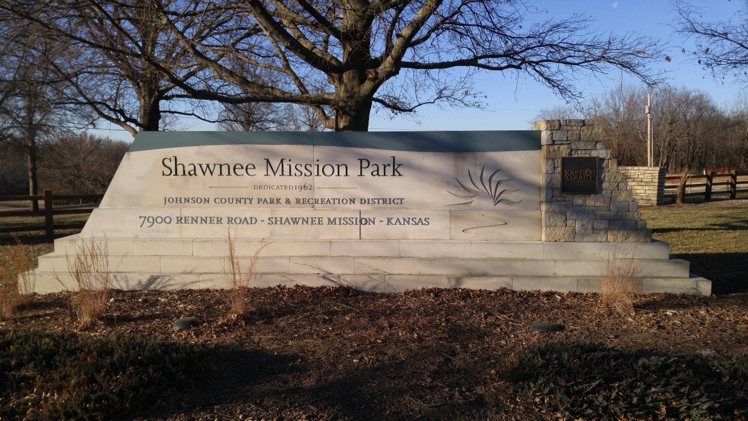 A stone monument sign for Shawnee Mission Park, located in Shawnee Mission, Kansas. The sign features the park's name, dedication year (1962), and additional information about the Johnson County Park & Recreation District. Surrounding the sign are patches of soil with dried grass and shrubs, and trees are visible in the background under a clear blue sky. Shawnee Mission Park mountain bike trail.